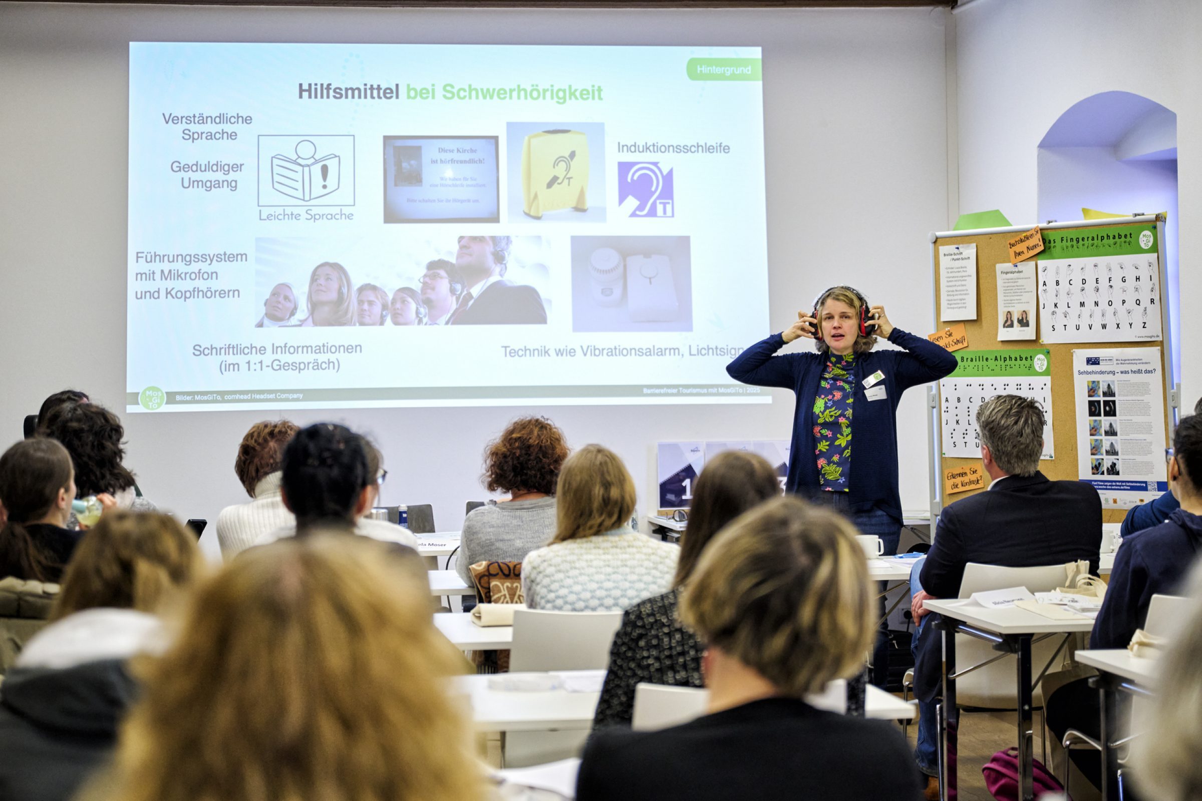 Referentin mit Kopfh&ouml;rern pr&auml;sentiert Folie zu Hilfsmitteln bei Schwerh&ouml;rigkeit vor Publikum im Seminarraum.
