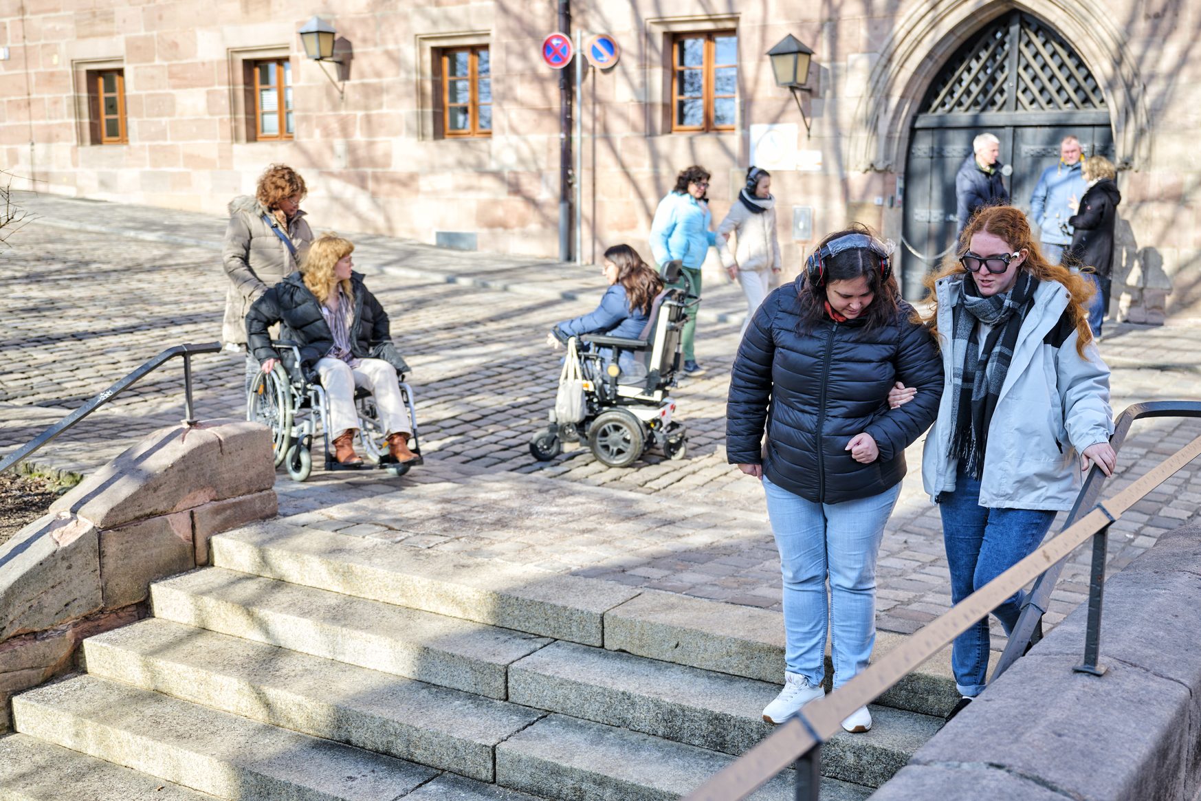 Menschen mit Behinderung im Au&szlig;enbereich vor einer historischen Fassade; zwei Frauen helfen am Treppenaufgang, weitere Personen im Hintergrund.