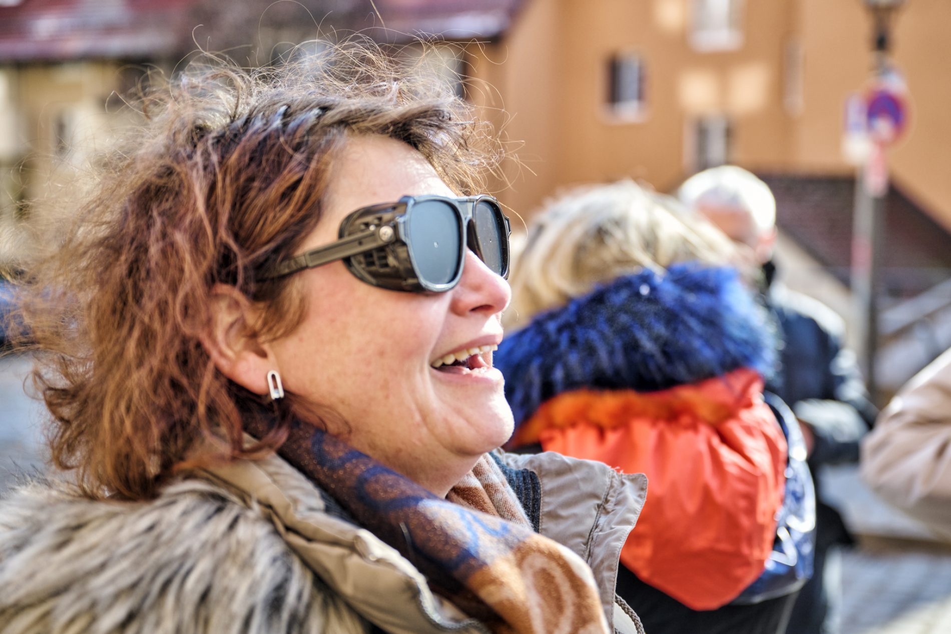 Sensibilisierungsübung Blindheit Frau mit Blindheits-Simulationsbrille lacht Outdoor, Jacke und Schal, Hintergrund mit weiteren Personen und Straßenzügen.
