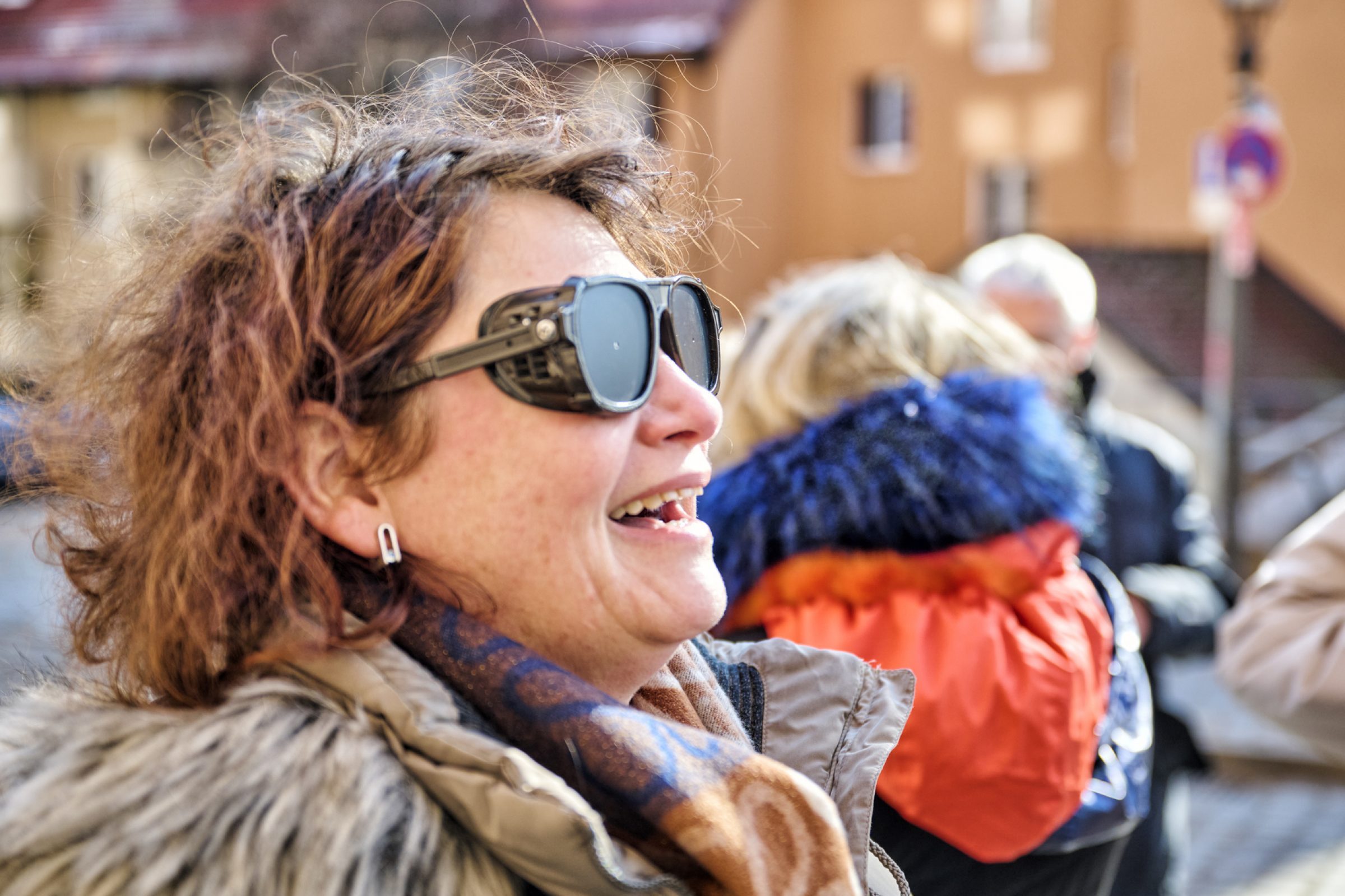 Frau mit Blindheits-Simulationsbrille lacht Outdoor, Jacke und Schal, Hintergrund mit weiteren Personen und Stra&szlig;enz&uuml;gen.