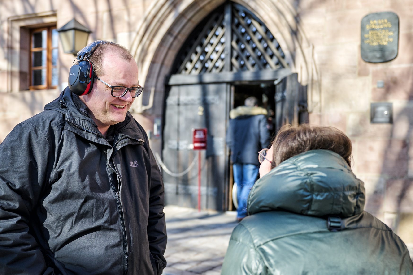 Mann mit Kopfh&ouml;rern l&auml;chelt im Gespr&auml;ch mit einer Person vor historischem Torbogen; Sensibilisierungs&uuml;bung Taubheit.