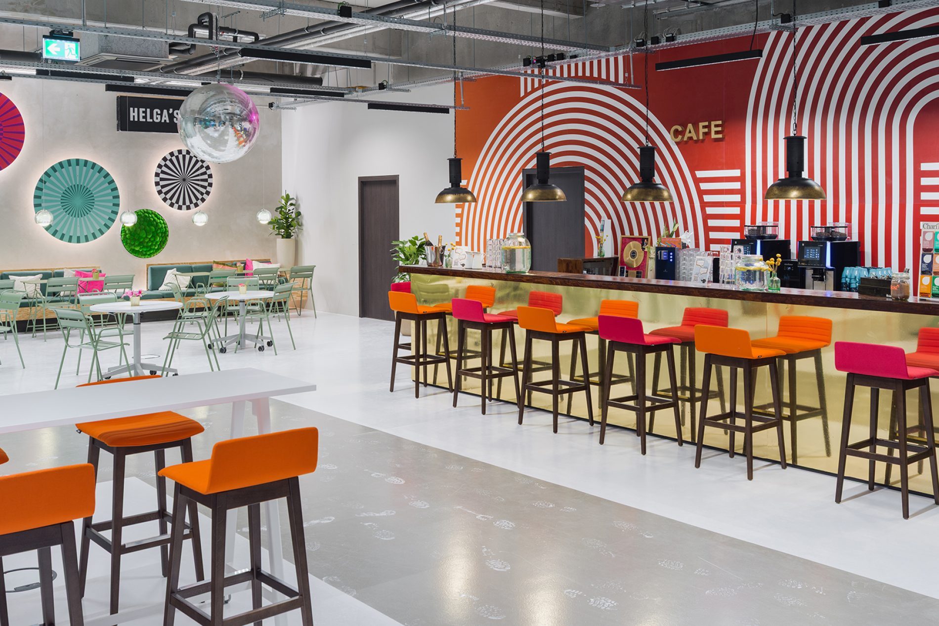 Heller Cafebereich mit langer Bar und bunten Hockern; kreisf&ouml;rmige Musterwand, Discokugel, Tische, Gr&uuml;npflanzen.