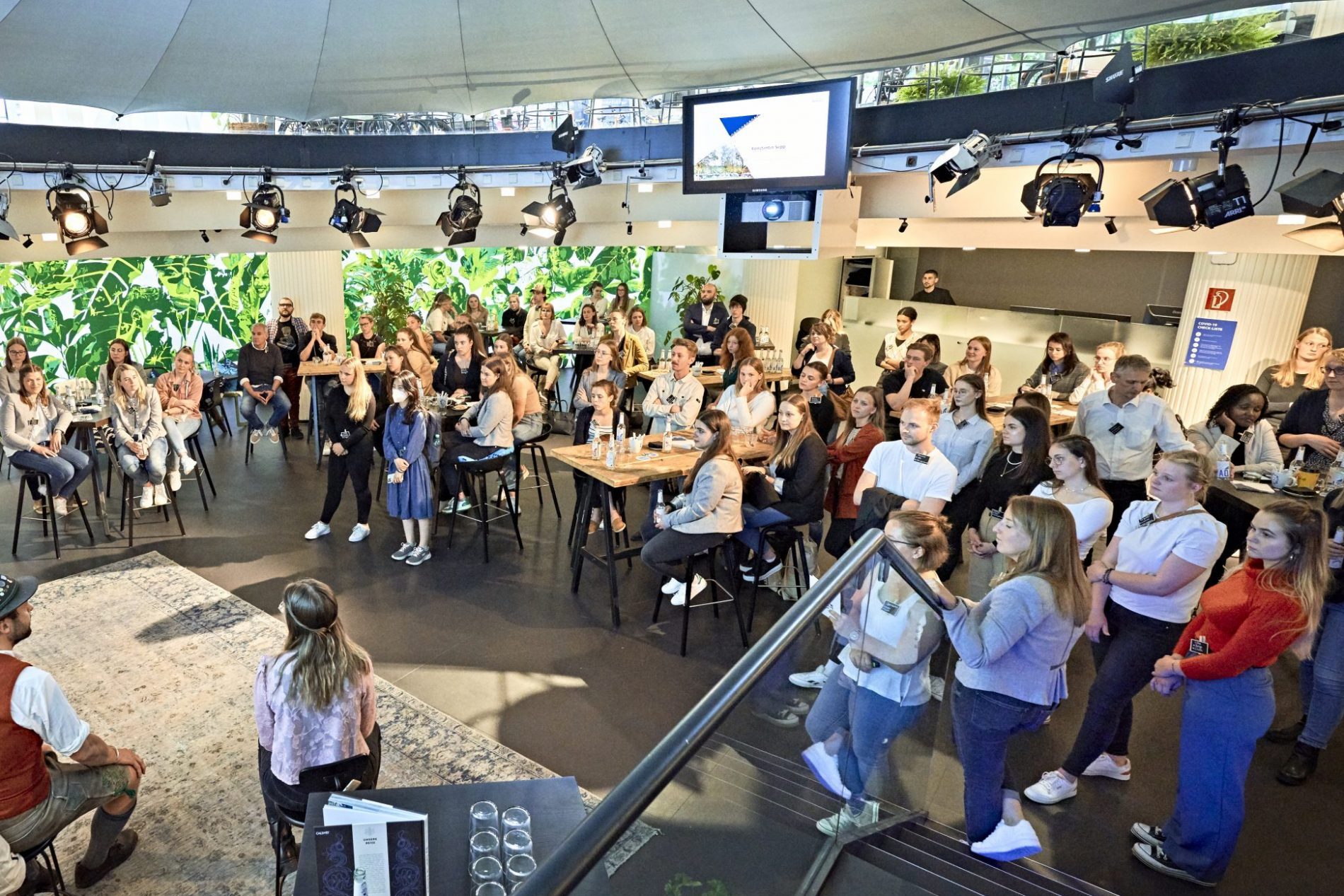 Gro&szlig;es Publikum sitzt an Tischen in moderner Halle mit gr&uuml;ner Pflanzenwand bei Tourism Next auf der ITB 2026.