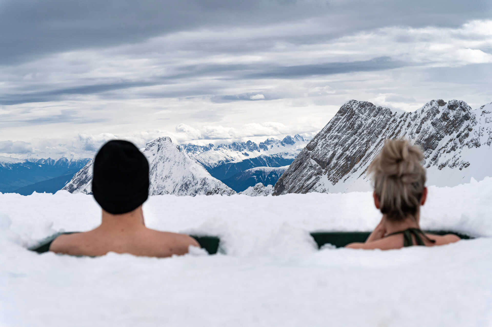 Zwei Personen sitzen in einem Eisloch, umgeben von Schnee, mit Blick auf die verschneiten Berge der bayerischen Alpen unter einem bewölkten Himmel.