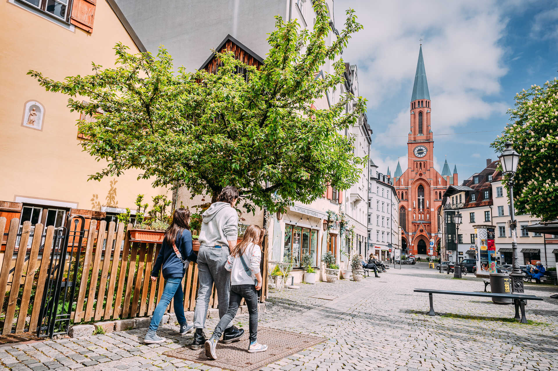 Familie spaziert durch eine sonnige Altstadtgasse mit Blick auf die rote Backsteinkirche St. Paul in München.