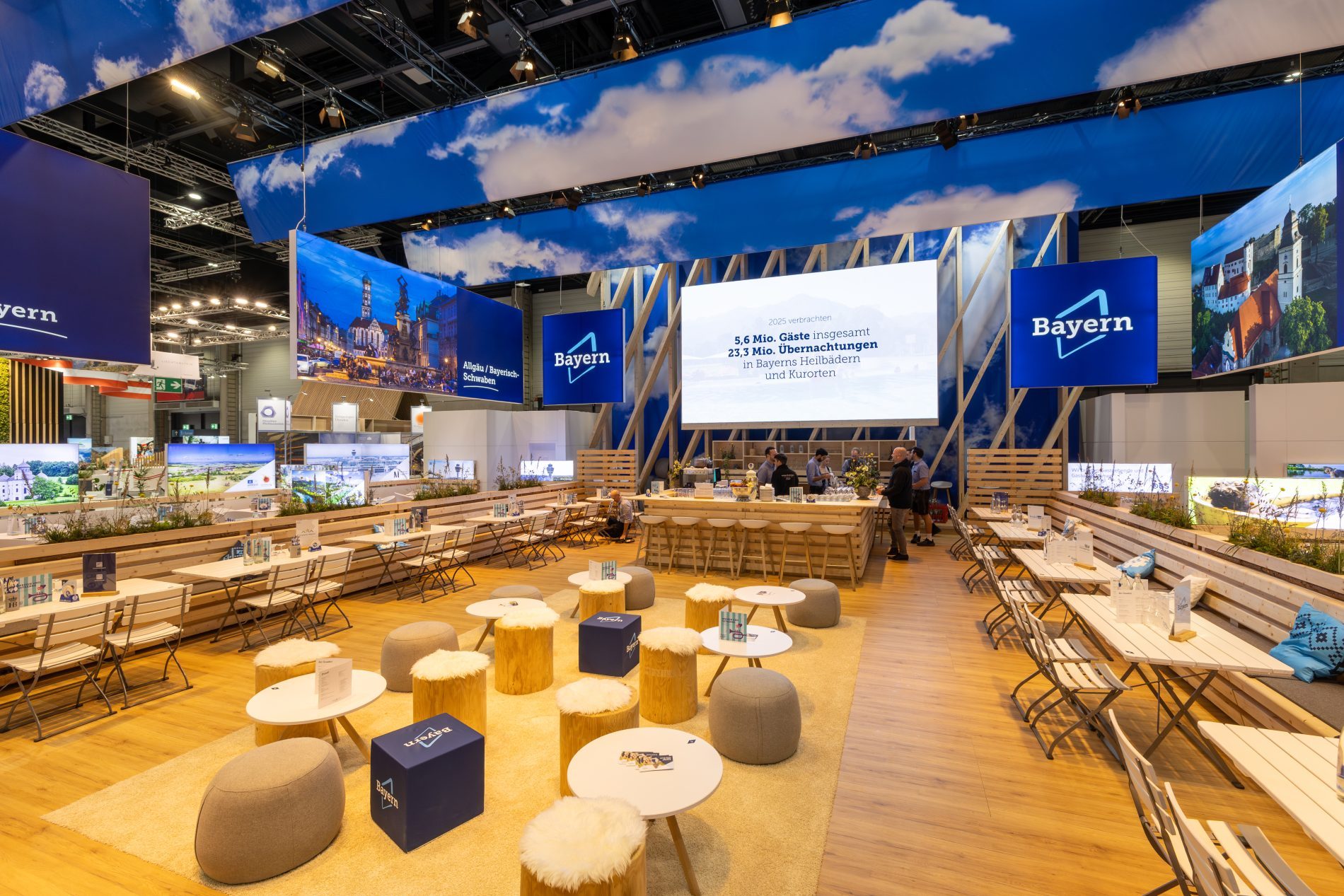 Bayern-Stand auf der ITB 2026 mit Sitzw&uuml;rfeln, Biergartentischen, Logo-Banner, gro&szlig;e LED-Wand und Bar.