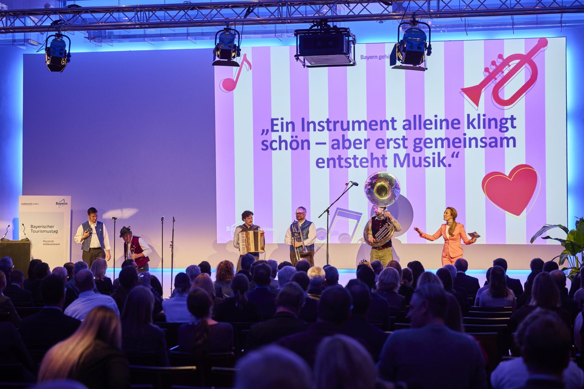 Musiker stehen auf einer Bühne während des Bayerischen Tourismustags; im Hintergrund eine große Leinwand mit farbigen Streifen und Musikinstrumenten, Publikum sitzt vor der Bühne.