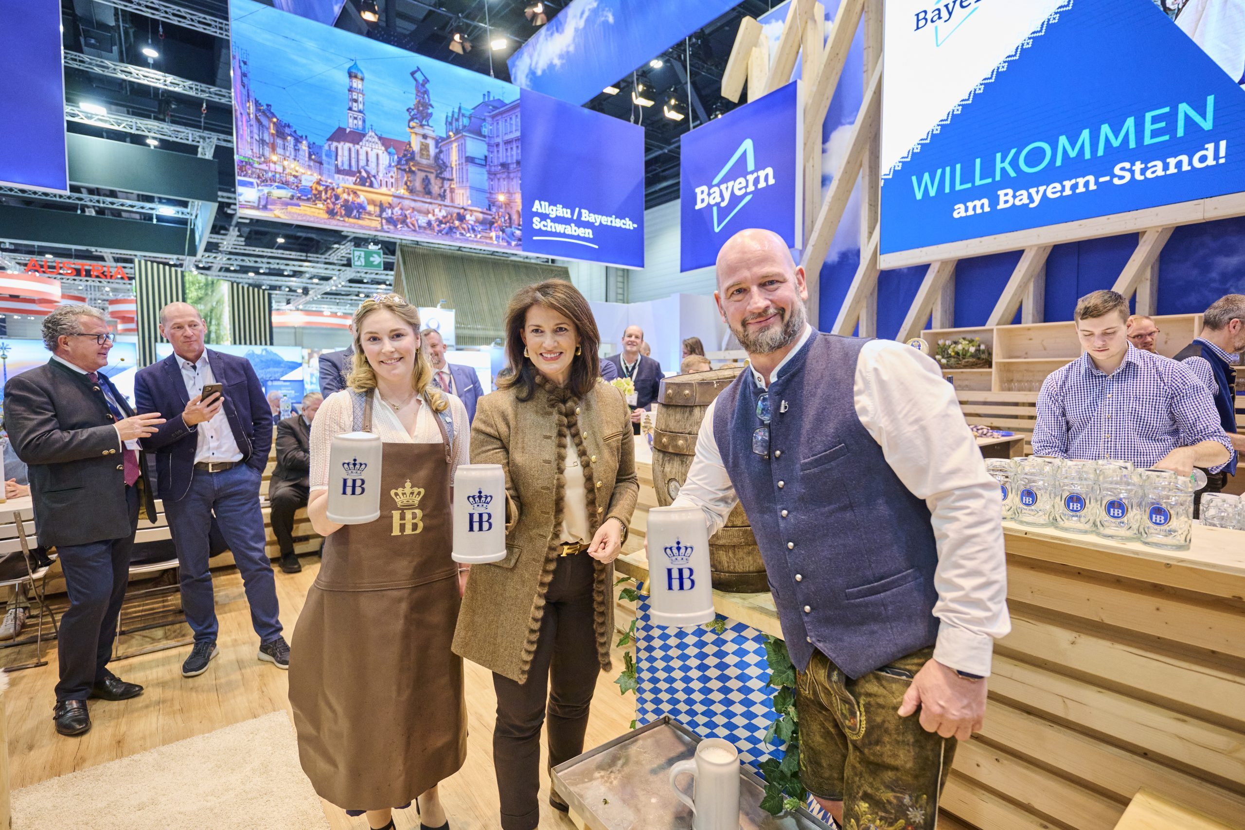 Bayern-Stand auf der ITB Berlin 2025; drei Personen pr&auml;sentieren Bierkr&uuml;ge mit HB-Logo vor bayerischer Dekoration und Besuchern.