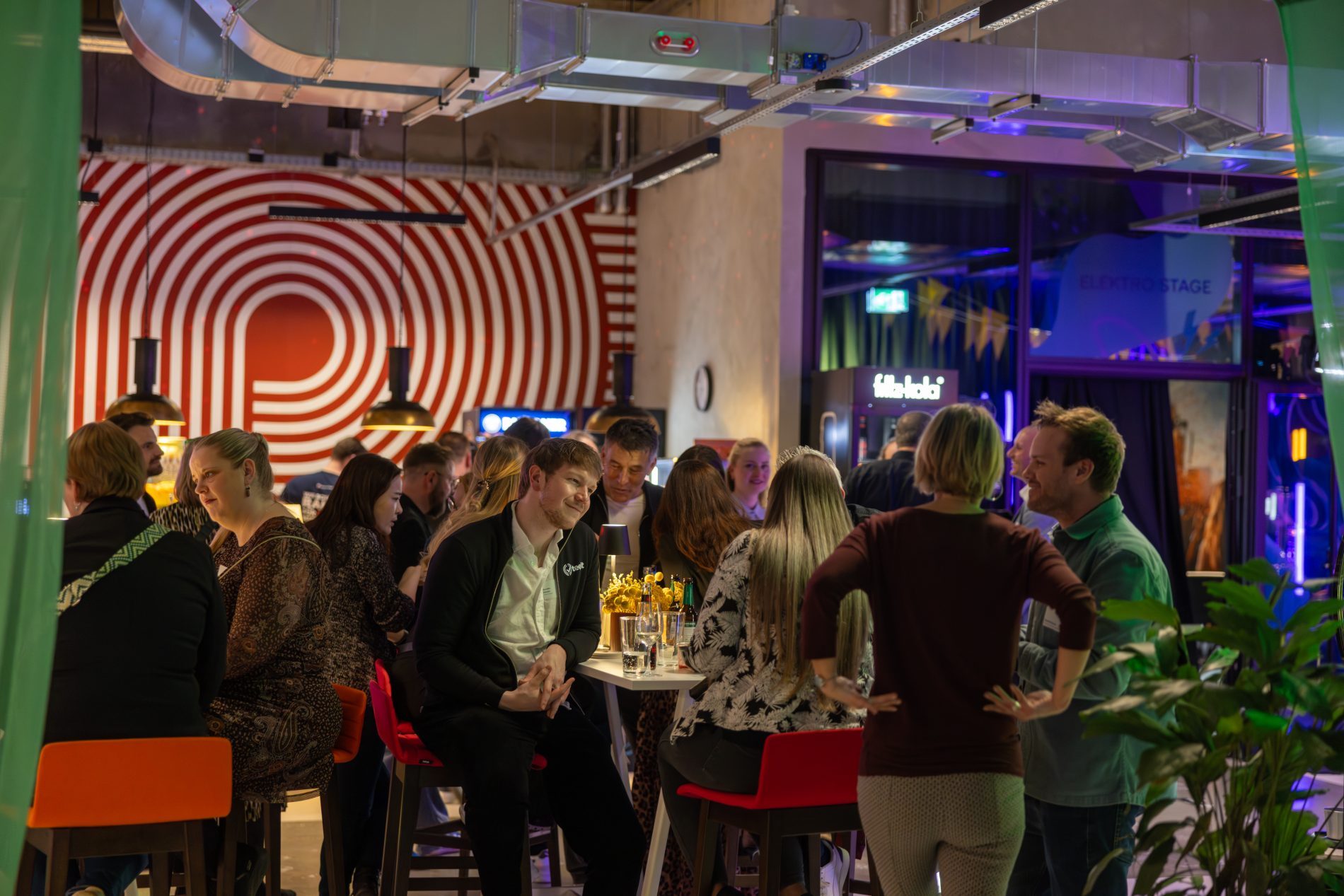 Gruppe von Teilnehmern an Stehtischen vor rot-wei&szlig;em Hintergrund und deckenhohem Fenster; gr&uuml;ne Gardinen und Pflanze im Vordergrund