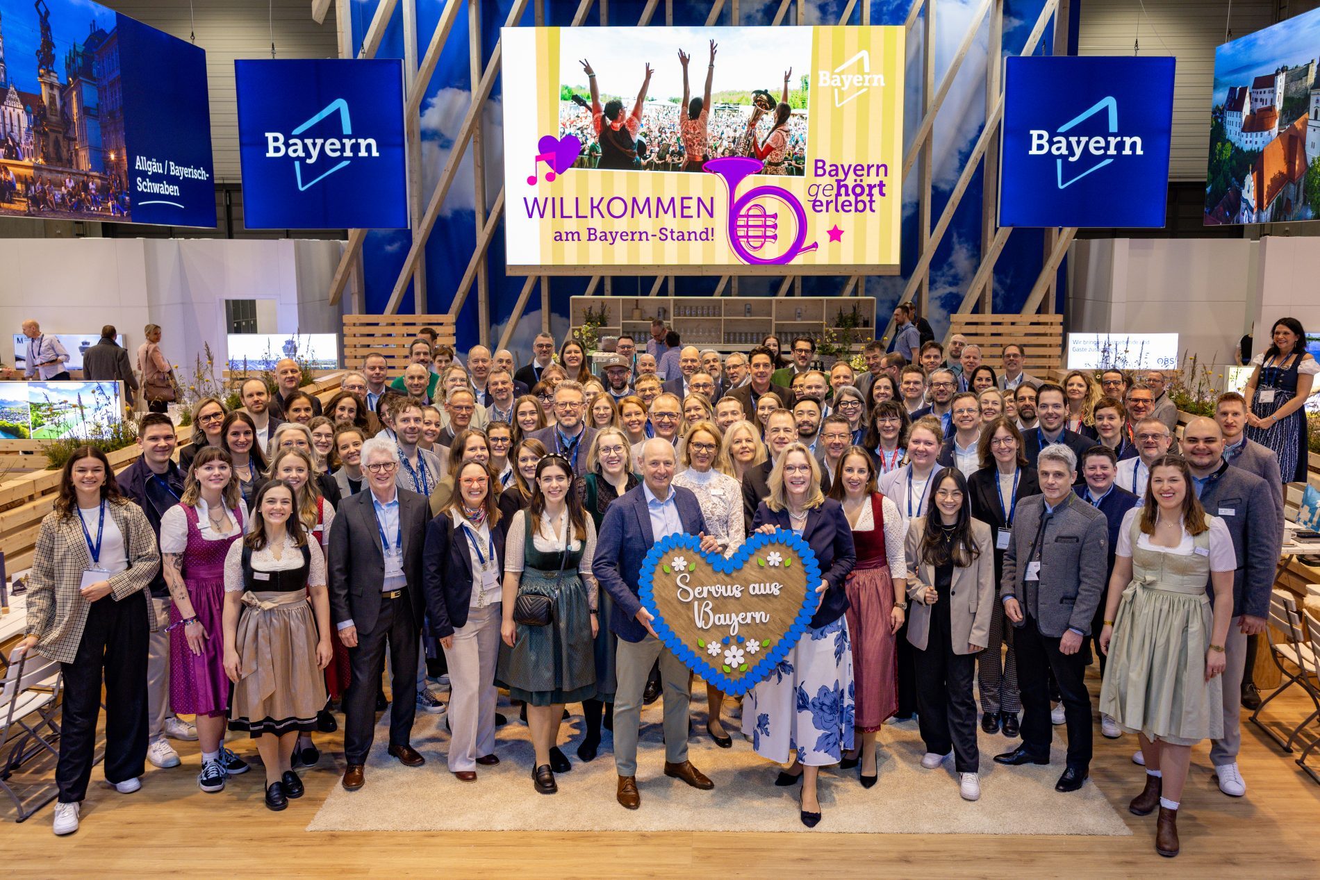 Gruppe von Teilnehmern vor gelbem Screen mit lila Schriftzug "Willkommen am Bayern Stand"; blau-wei&szlig;es Bayern Logo links und rechts von Screen, 2 Personen im Vordergrund halten gro&szlig;es Lebkuchenherz aus Holz mit Schriftzug "Servus aus Bayern" in der Hand