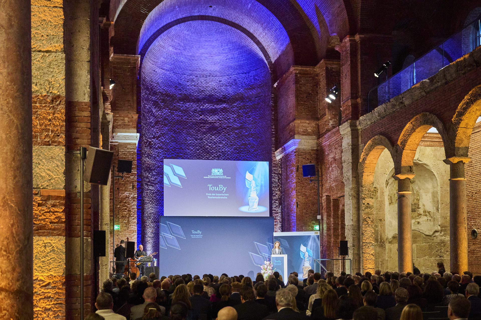 Eine Veranstaltung im historischen Geb&auml;ude mit einem Redner und Publikum, umgeben von bunten Lichtern und einer gro&szlig;en Pr&auml;sentationsleinwand.