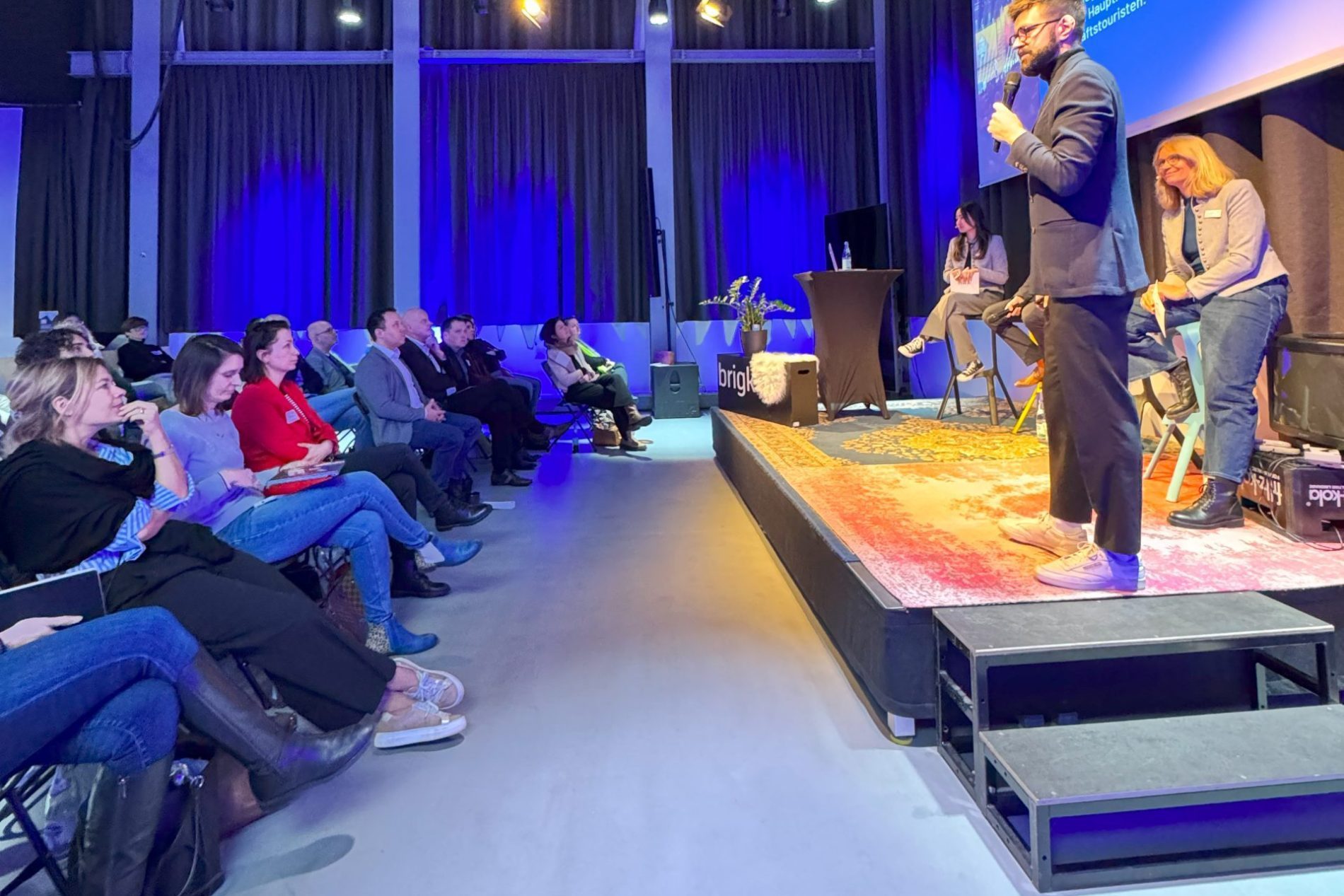 Keynote Speaker h&auml;lt Rede auf einer B&uuml;hne vor Publikum. Im Hintergrund ist ein Bildschirm mit einer Pr&auml;sentation zu sehen.