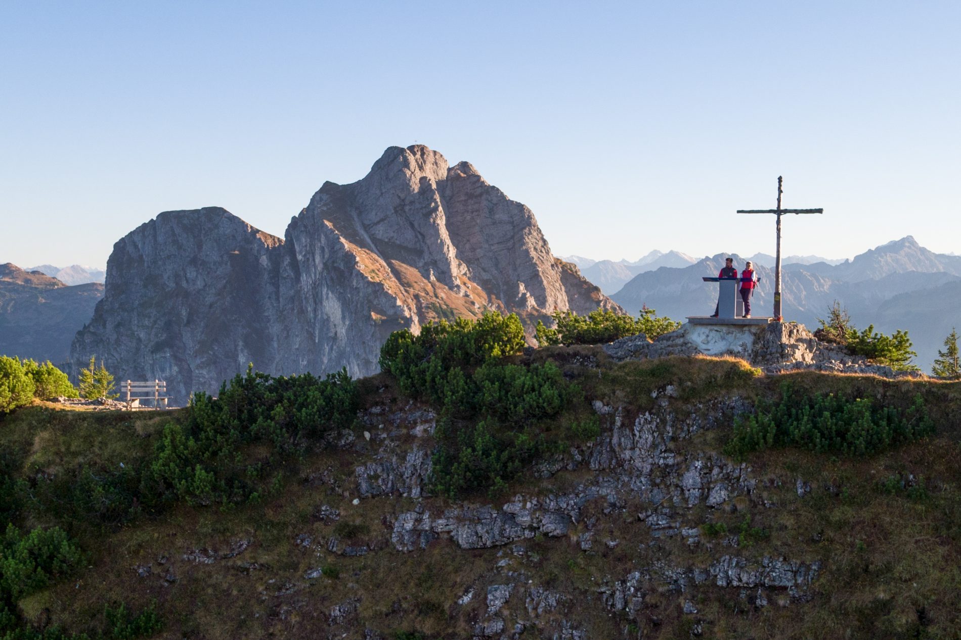 Paar steht in Wanderkleidung neben einem Gipfelkreuz, im Hintergrund ist das Bergpanorama zu sehen.