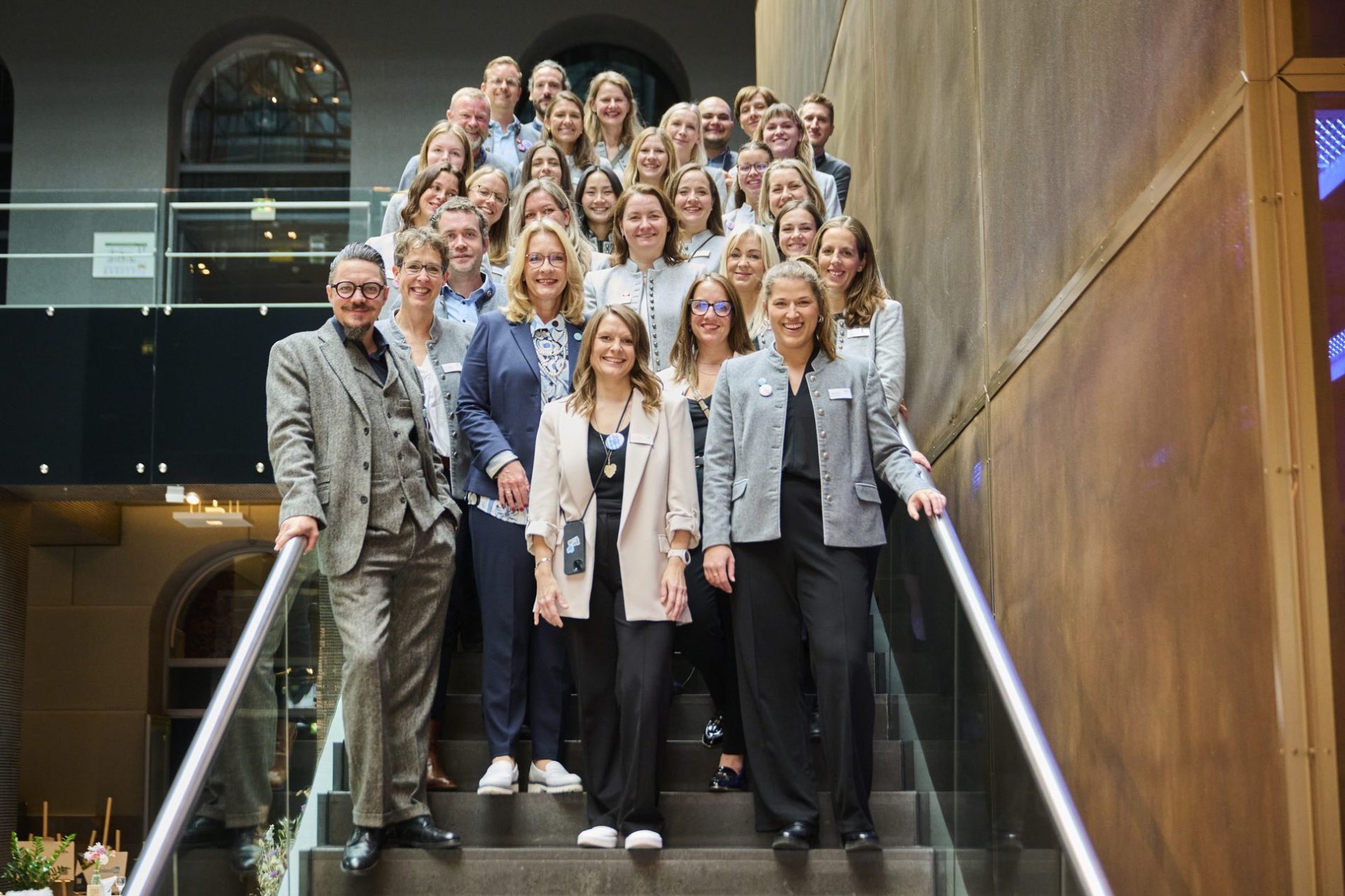 Gruppe von ca. 30 Personen auf einer Treppe, l&auml;chelnd, in formeller Kleidung, im Innenbereich eines Geb&auml;udes.