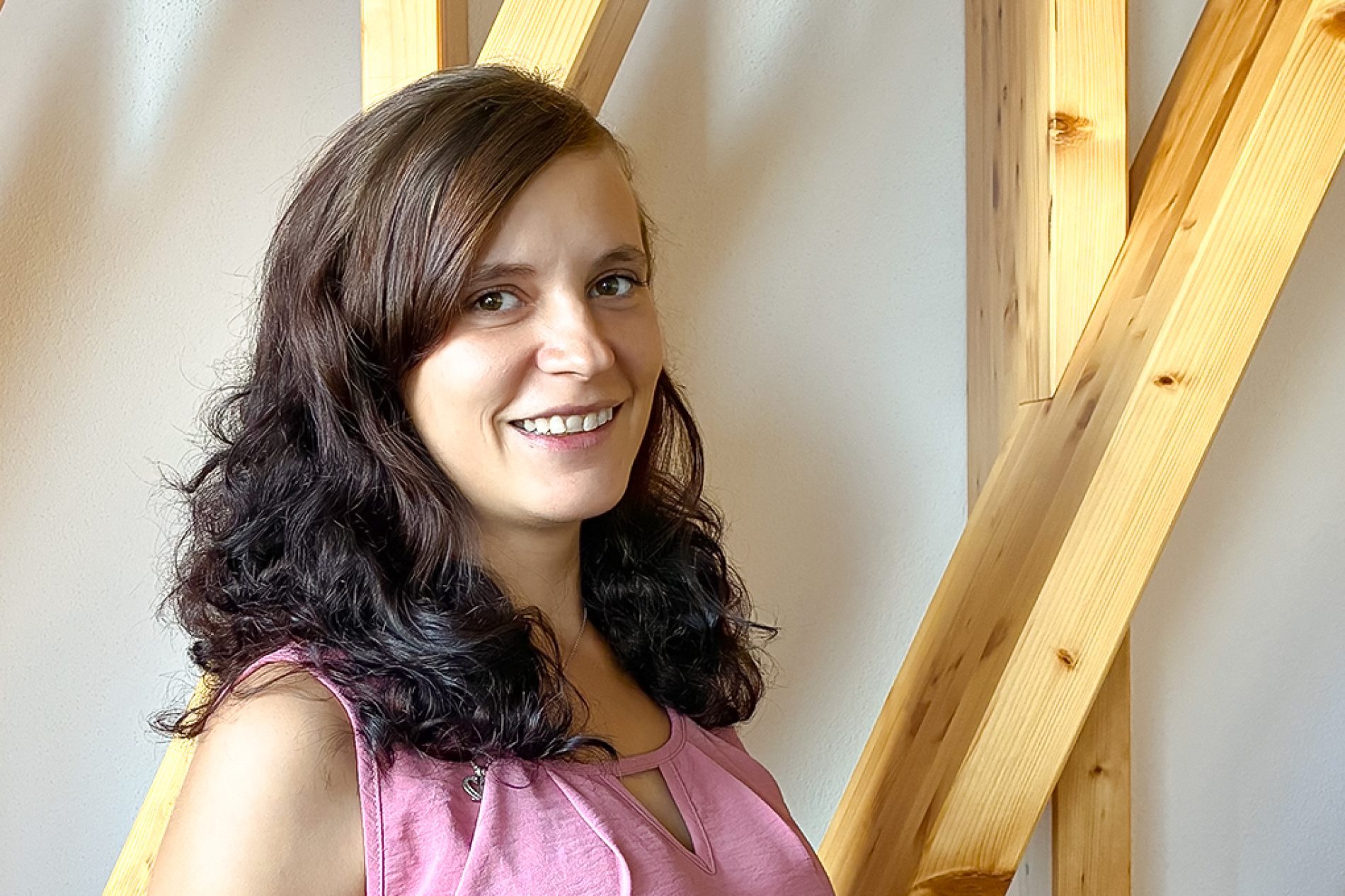 Frau mit langen, lockigen Haaren l&auml;chelt vor einer Wand mit Holzelementen. Sie tr&auml;gt ein rosa Oberteil.